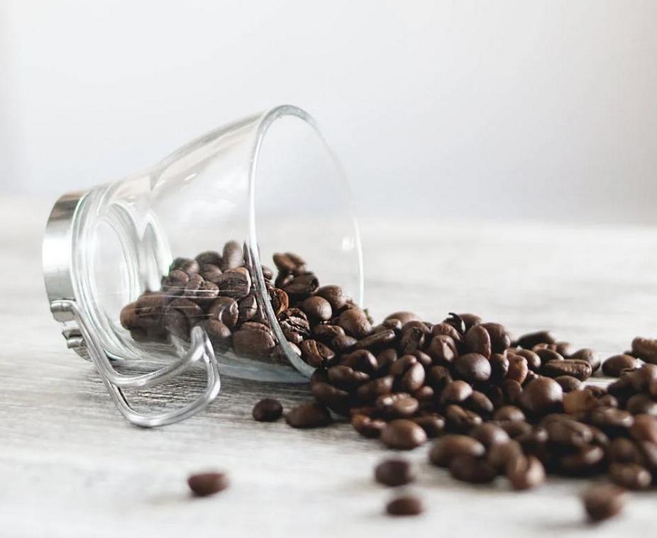 A clear glass cup tipped over with coffee beans spilled onto a wooden surface.