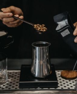 A person measuring coffee beans with a metal scoop over a metallic jug on a digital kitchen scale.