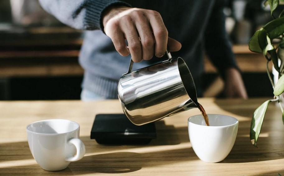 A person pouring hot water from a metal kettle into a white ceramic cup on a wooden table with a smartphone and plant in the background.