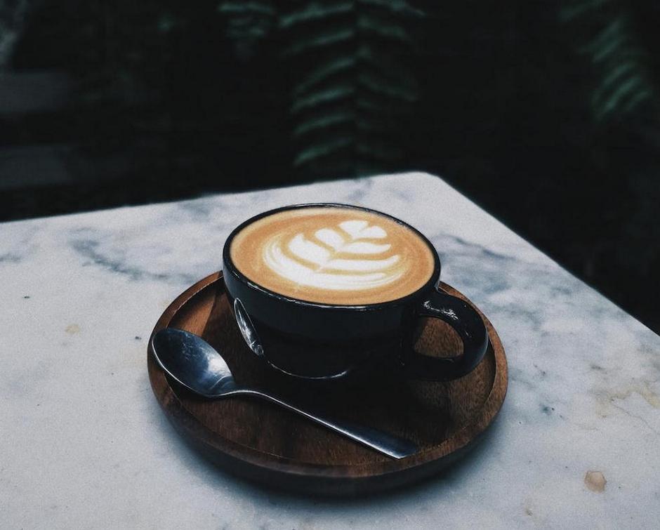 A latte with artful foam design in a black cup on a wooden saucer with a spoon, placed on a marble surface with ferns in the background.